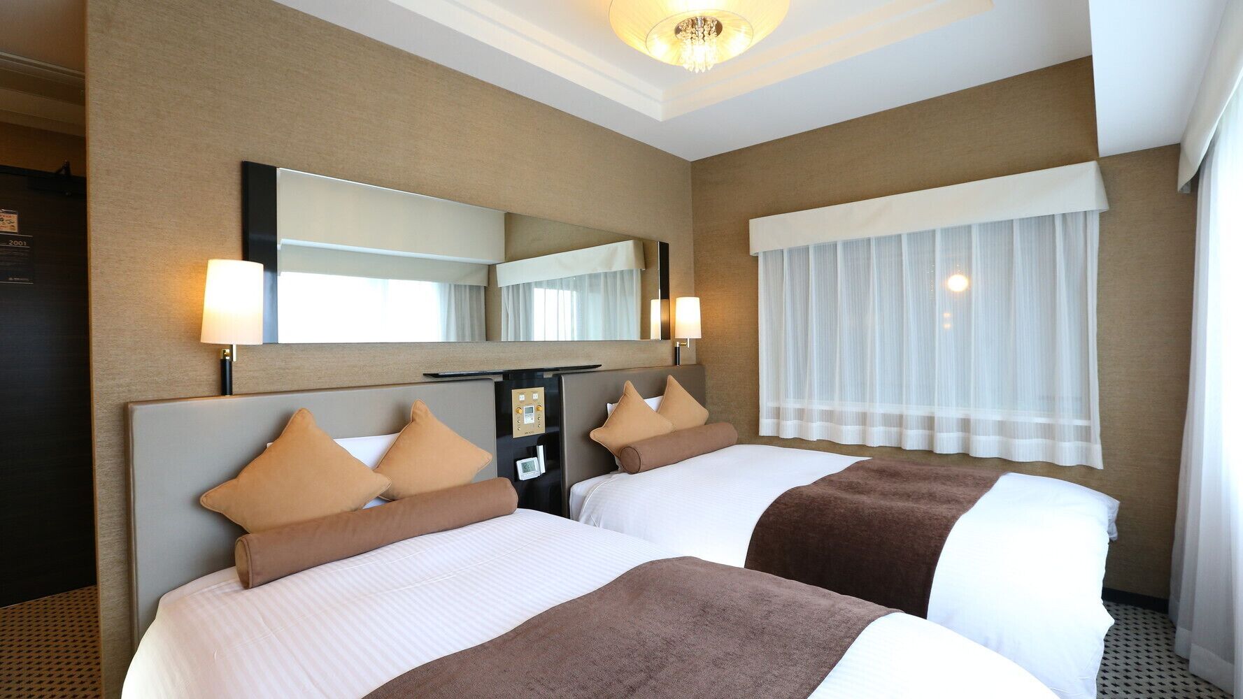 Een hotelkamer met twee bedden, een grote spiegel, een bedieningspaneel en een raam met dunne gordijnen.