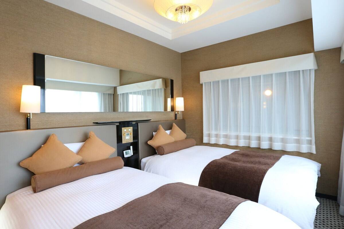 Habitación de hotel con dos camas, un espejo grande, un panel de control y una ventana con cortinas translúcidas.