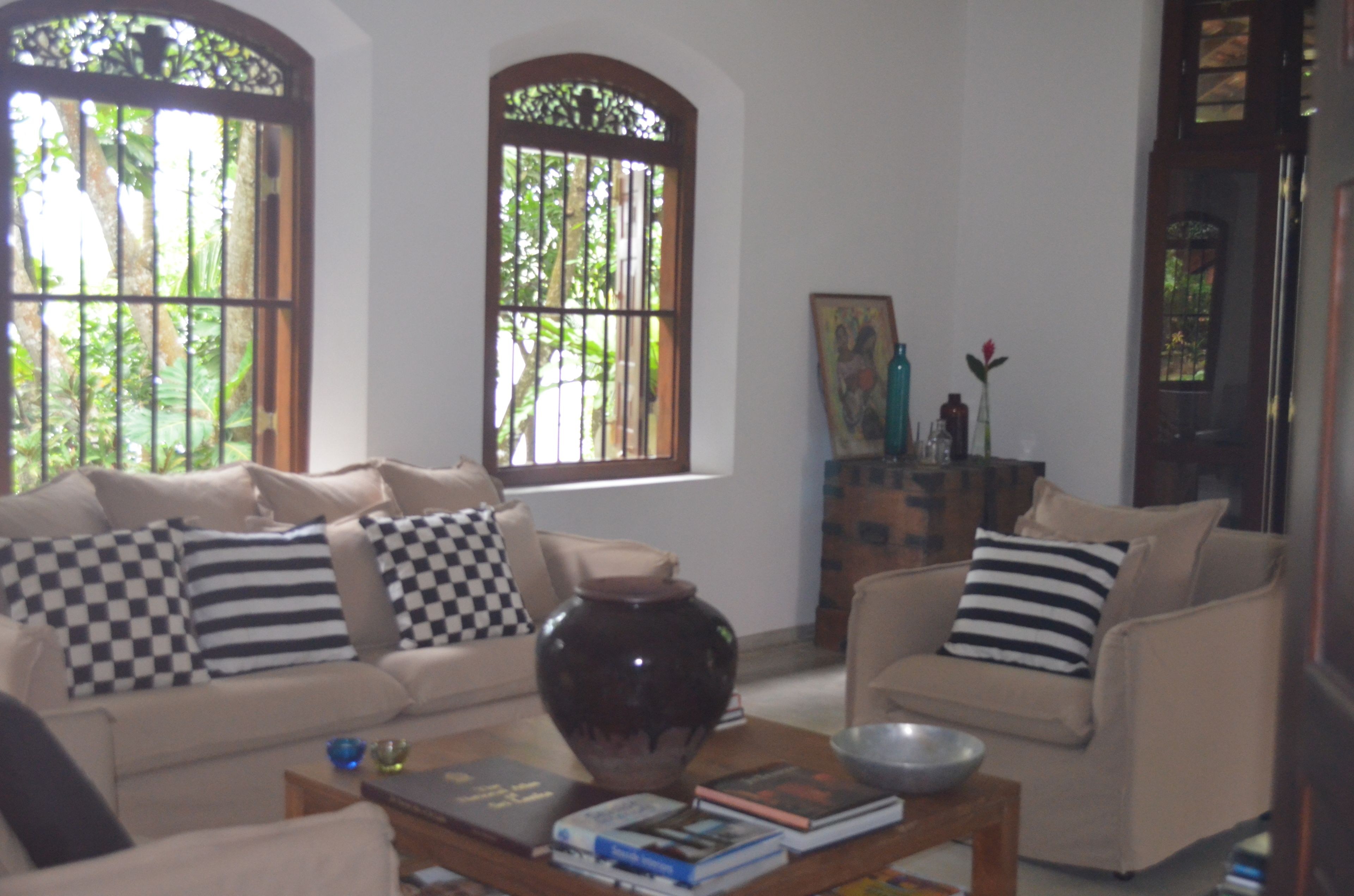 Una sala de estar con un sofá color crema, una mesa baja con libros, un jarrón grande y dos ventanas con rejas.