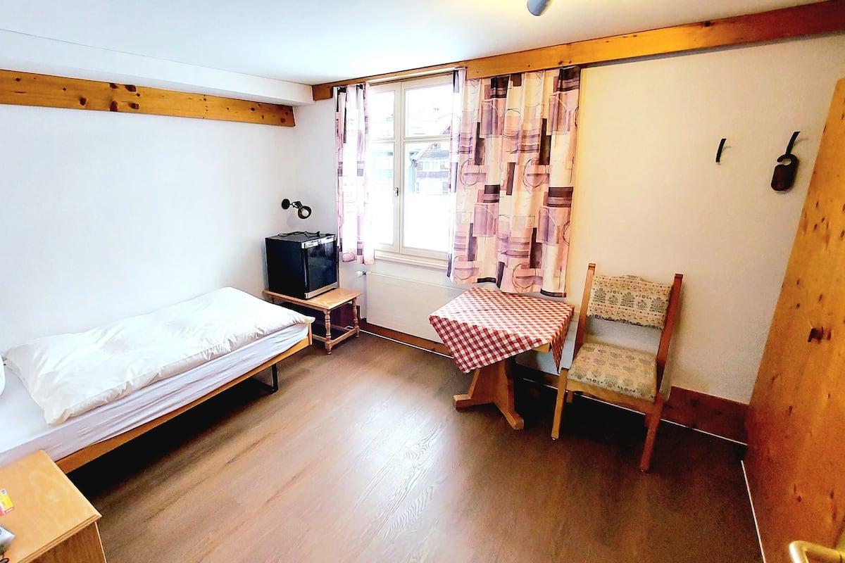 Um quarto pequeno e acolhedor com uma cama de solteiro, uma mesa de madeira, uma cadeira, uma televisão e uma janela com cortinas.