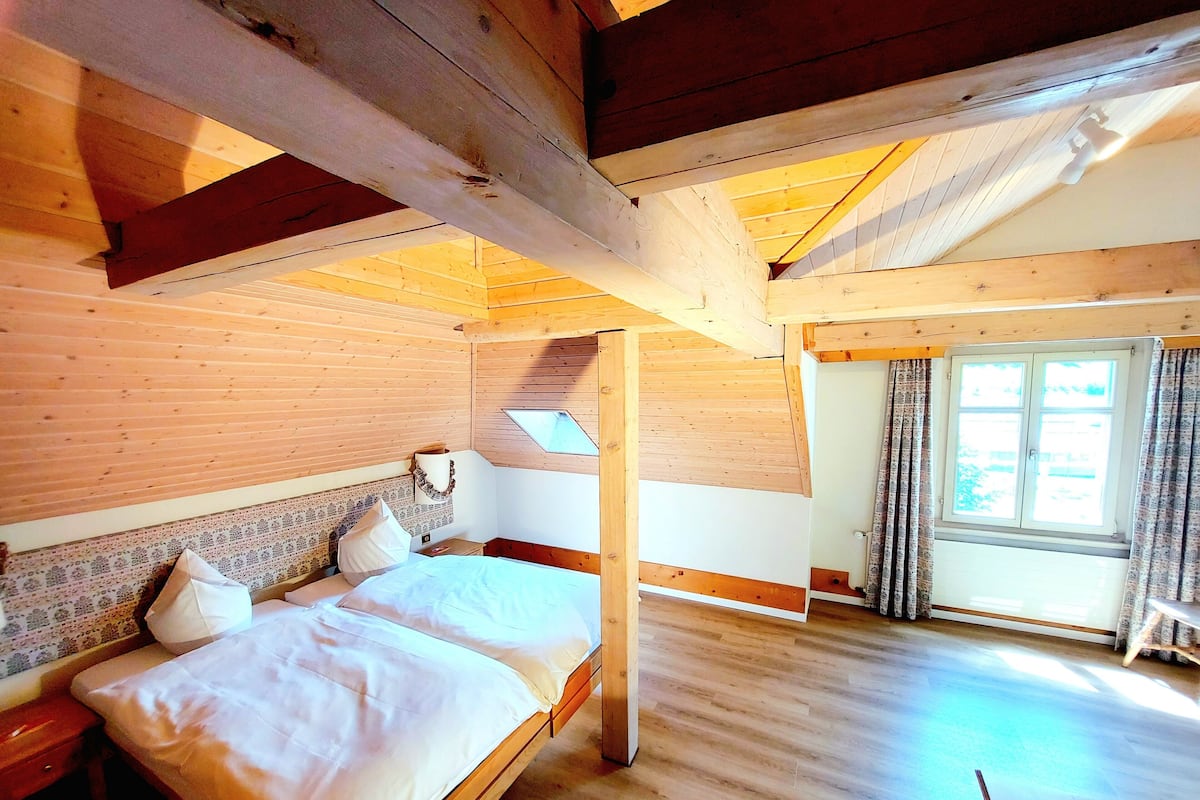 Quarto de madeira com cama, mesa de cabeceira, janela com cortinas e teto inclinado.