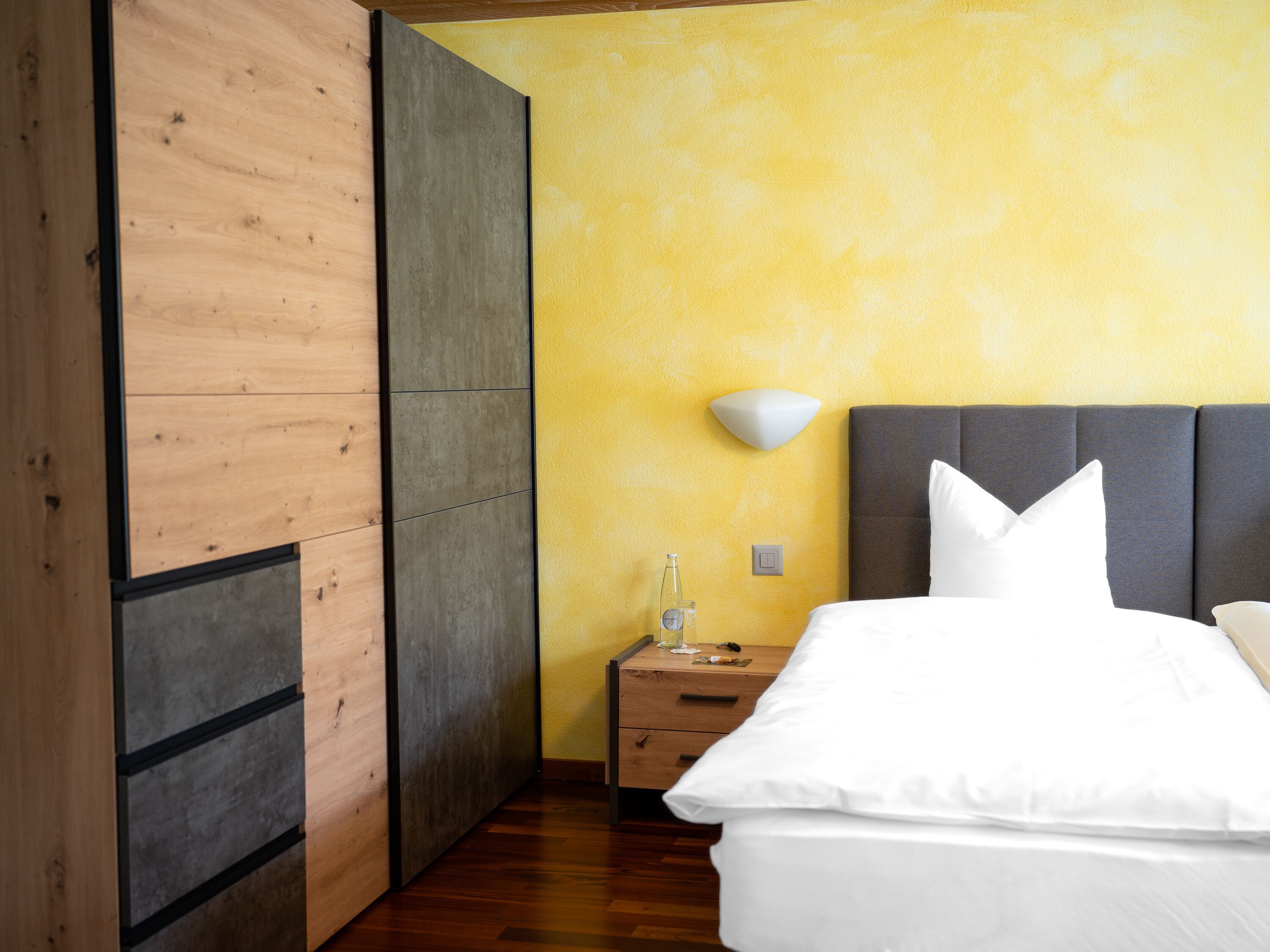 A modern bedroom with a wooden wardrobe, a bedside table with a bottle, and a bed with white linens.