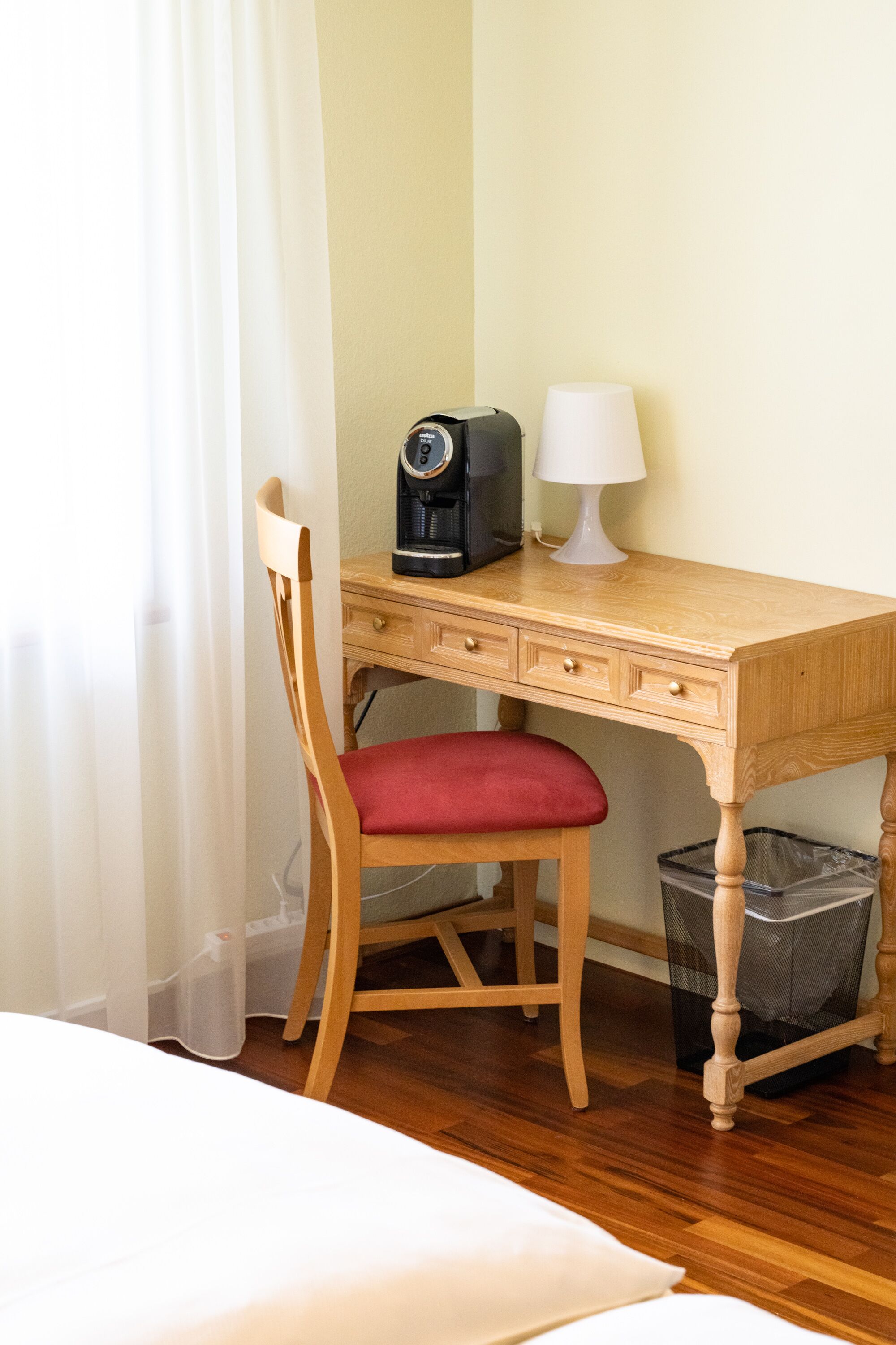 A wooden desk with a red chair, a coffee maker, and a lamp.