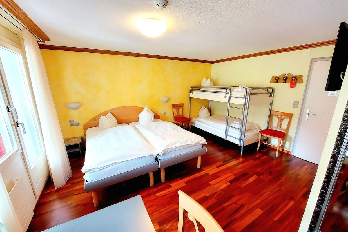 Quarto com duas camas, uma mesa e uma cadeira. Há uma luminária no teto e uma porta.