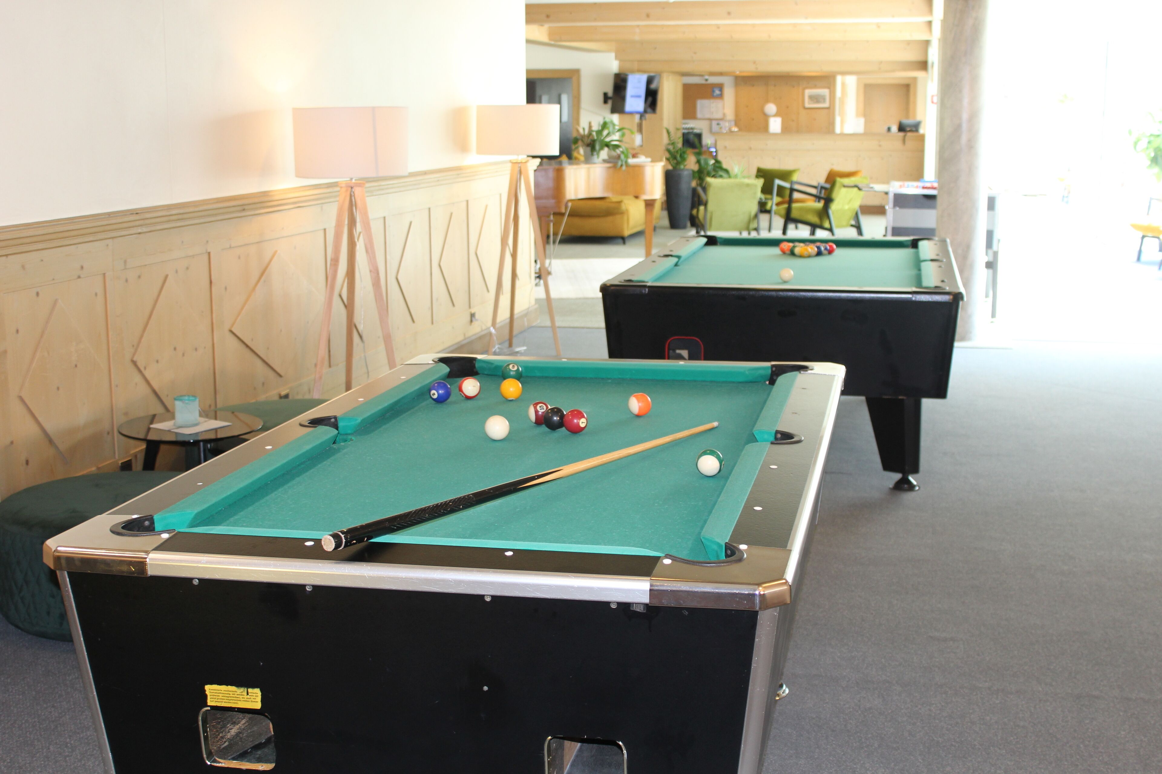Une table de billard avec des billes et une queue de billard, une deuxième table en arrière-plan, et un espace d’accueil avec un téléviseur et des plantes.