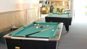 Une table de billard avec des billes et une queue de billard, une deuxième table en arrière-plan, et un espace d’accueil avec un téléviseur et des plantes.
