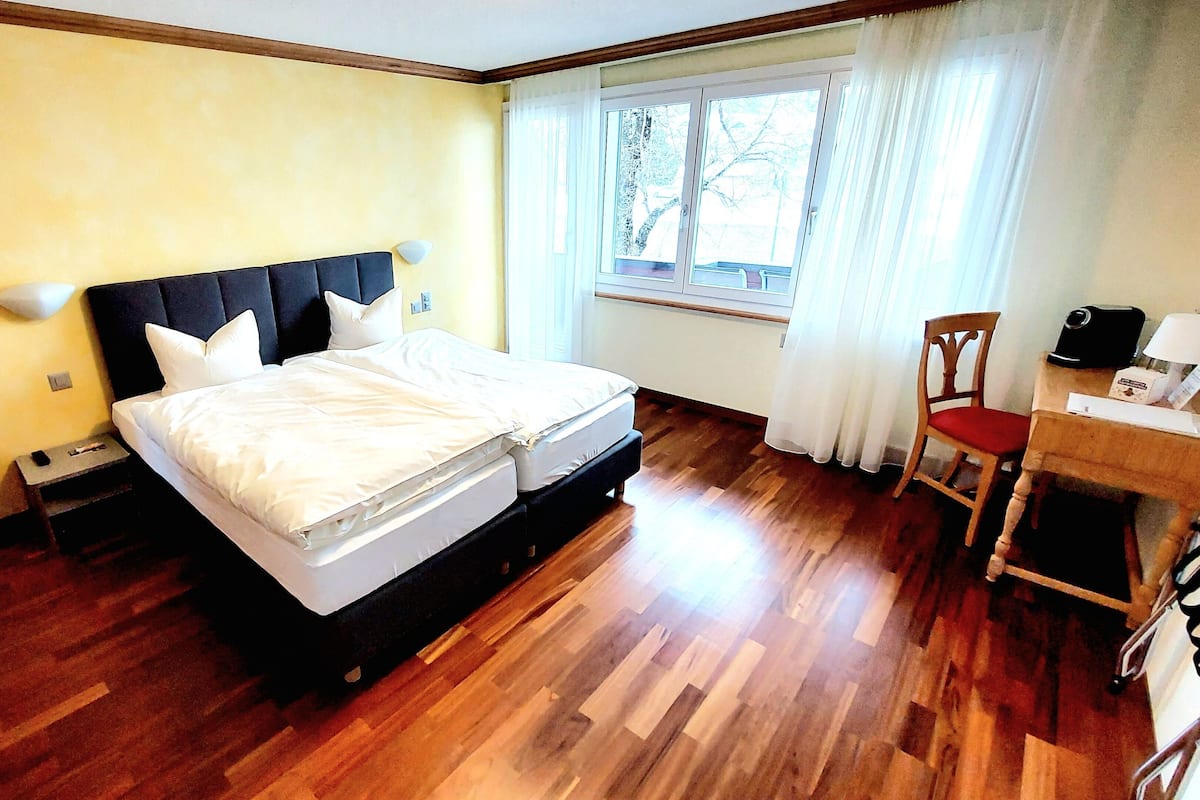 Quarto de hotel com cama de casal, piso de madeira, escrivaninha com cadeira e janela com cortinas.