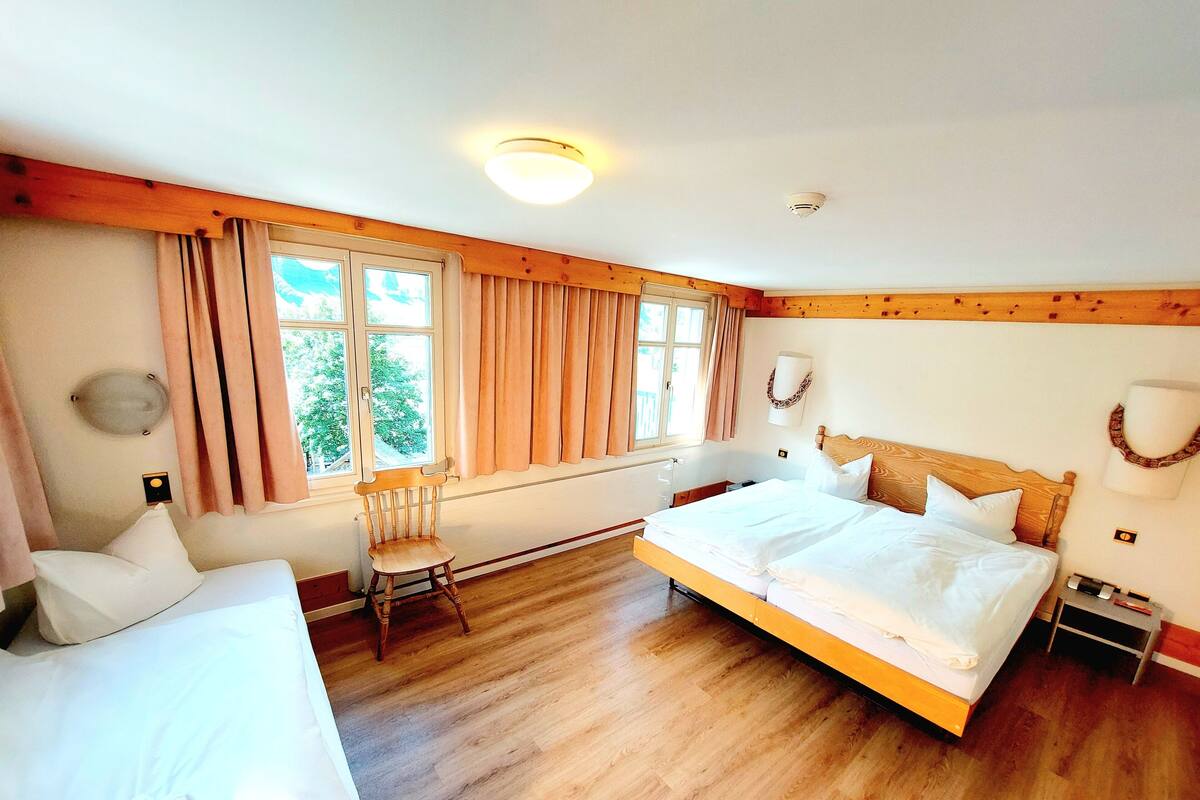 Quarto de hotel com uma cama grande, uma cama menor, uma cadeira, uma janela com cortinas e uma luminária no teto.