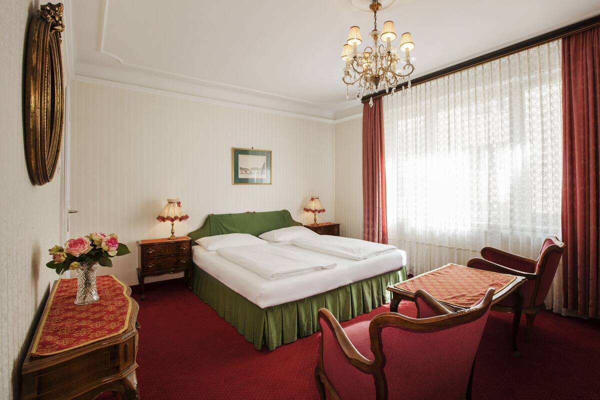 A hotel room with a double bed, two bedside tables with lamps, a green upholstered headboard, a wooden chair, a table with a red tablecloth, a chandelier, and a window with sheer curtains.
