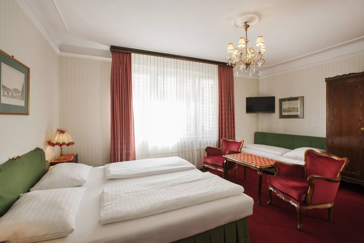 A hotel room with two beds, a green sofa, a red armchair, a small table, and a chandelier.