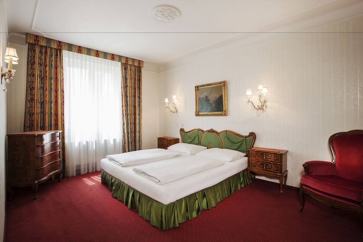 A hotel room with a large bed, two bedside tables, a red armchair, a wooden dresser, and a painting on the wall.