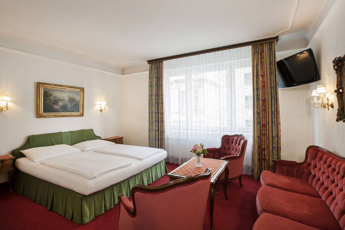 A hotel room with a double bed, a small table with a vase, a red armchair, a red sofa, a television, and a painting on the wall.
