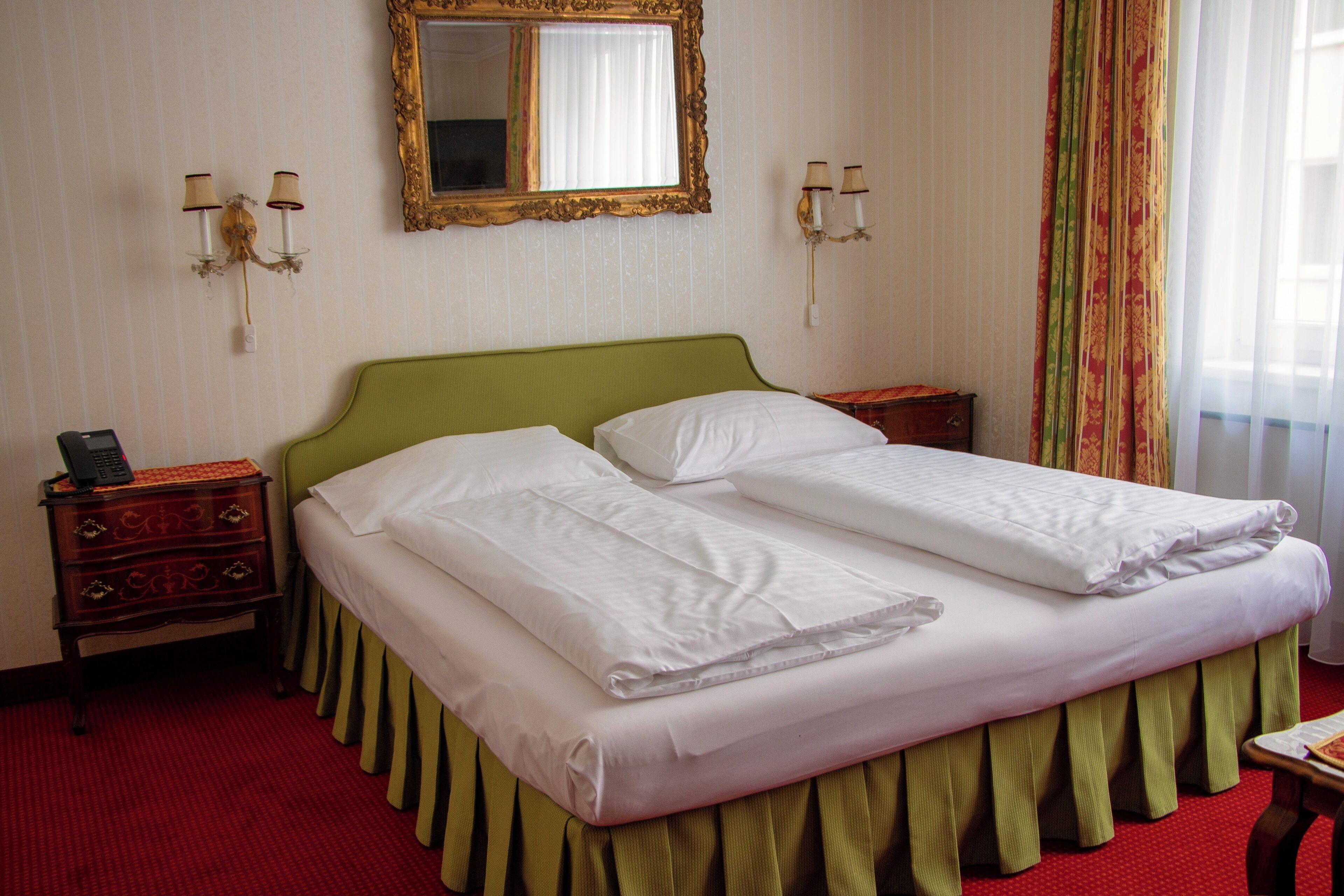 A hotel room with a green headboard, two white beds, a wooden nightstand with a telephone, and a mirror on the wall.
