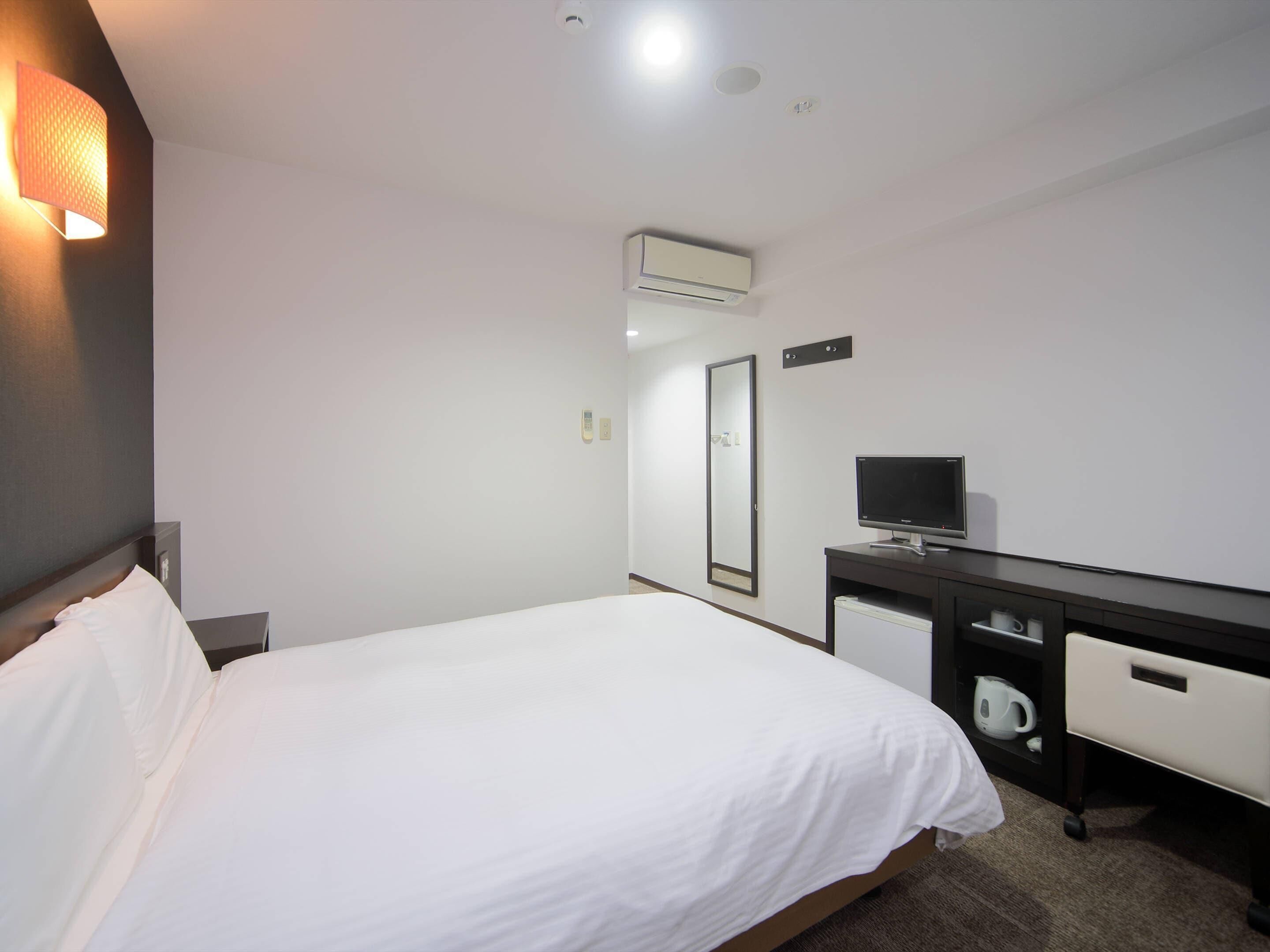 A hotel room with a bed, a desk with a computer, a mirror, and an air conditioning unit.