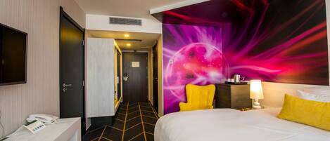 A hotel room with a bed, a desk with a telephone, a chair, and a wall with a vibrant abstract design.