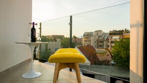 Un balcon avec un tabouret jaune, une bouteille de vin et un verre posés sur une petite table blanche.