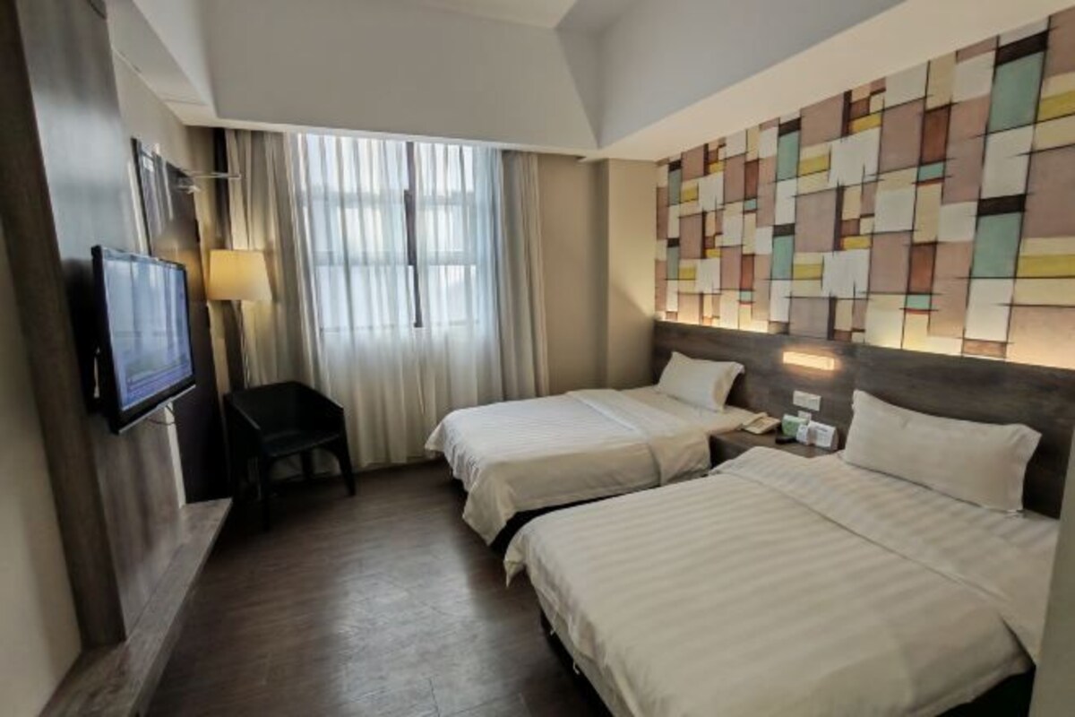 A hotel room with two beds, a TV mounted on the wall, a desk, and a window with curtains.