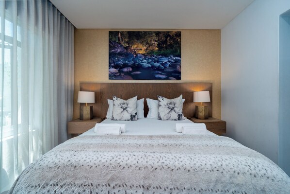 A hotel room with a large bed, two bedside lamps, a painting of a forest stream, and a view of the outdoors.