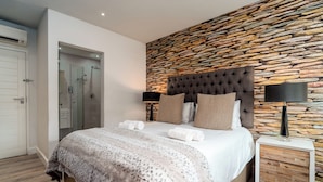A bedroom with a stone wall, a tufted headboard, a bedside table with a lamp, and a bed with a patterned blanket.
