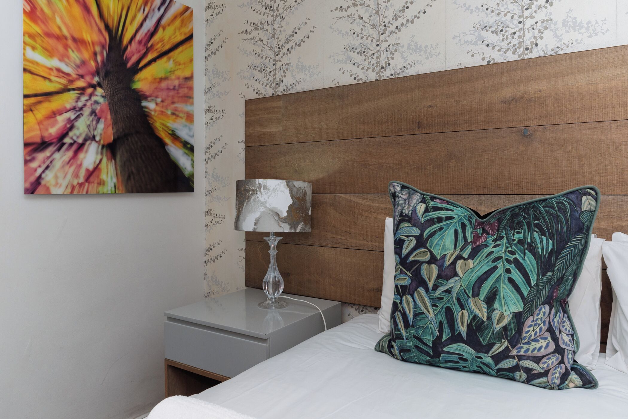 Una cama bien tendida con un cojín floral, una lámpara de noche y una pared con estampado floral.