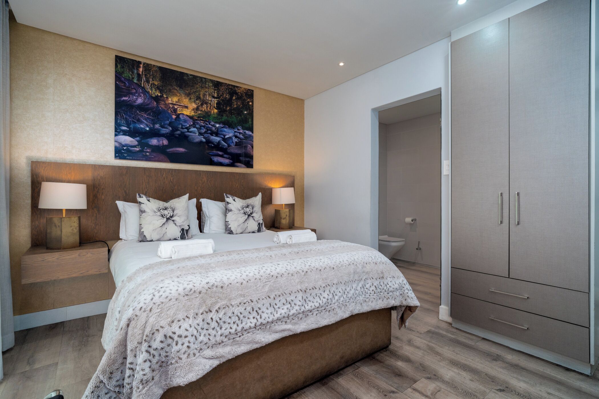A modern bedroom with a large bed, bedside lamps, a painting above the bed, and a wardrobe.