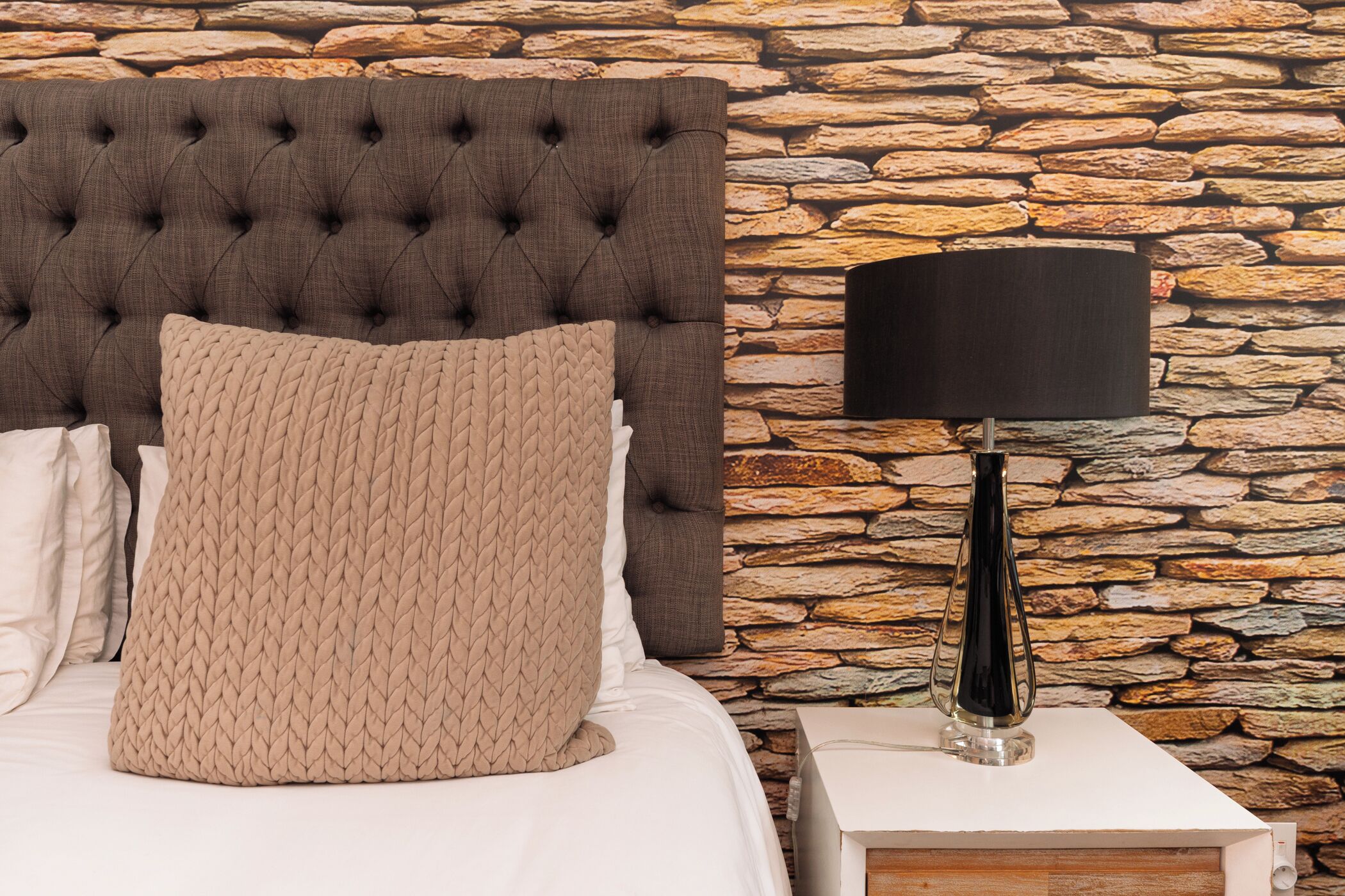 Una cama con cabecera capitonada, un cojín tejido color beige y una mesita de noche con una lámpara negra.
