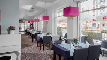 Un restaurant avec des tables dressées, de grandes fenêtres et des lampes suspendues roses.