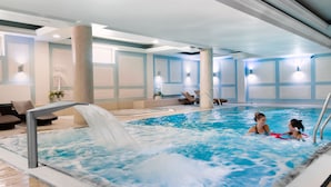 Una piscina interior con dos personas nadando, sillas de descanso y una fuente.