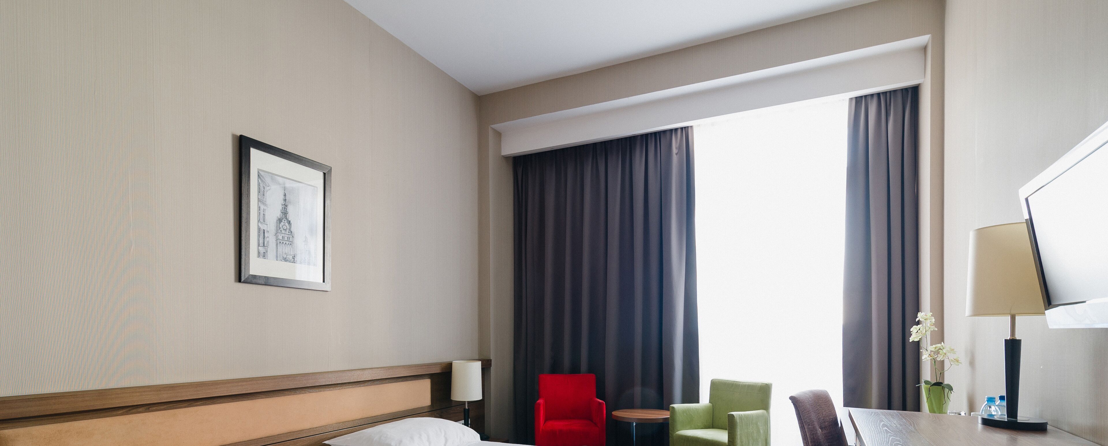 A hotel room with a bed, a framed picture on the wall, a red chair, a green chair, a small table, a lamp, and a window with curtains.