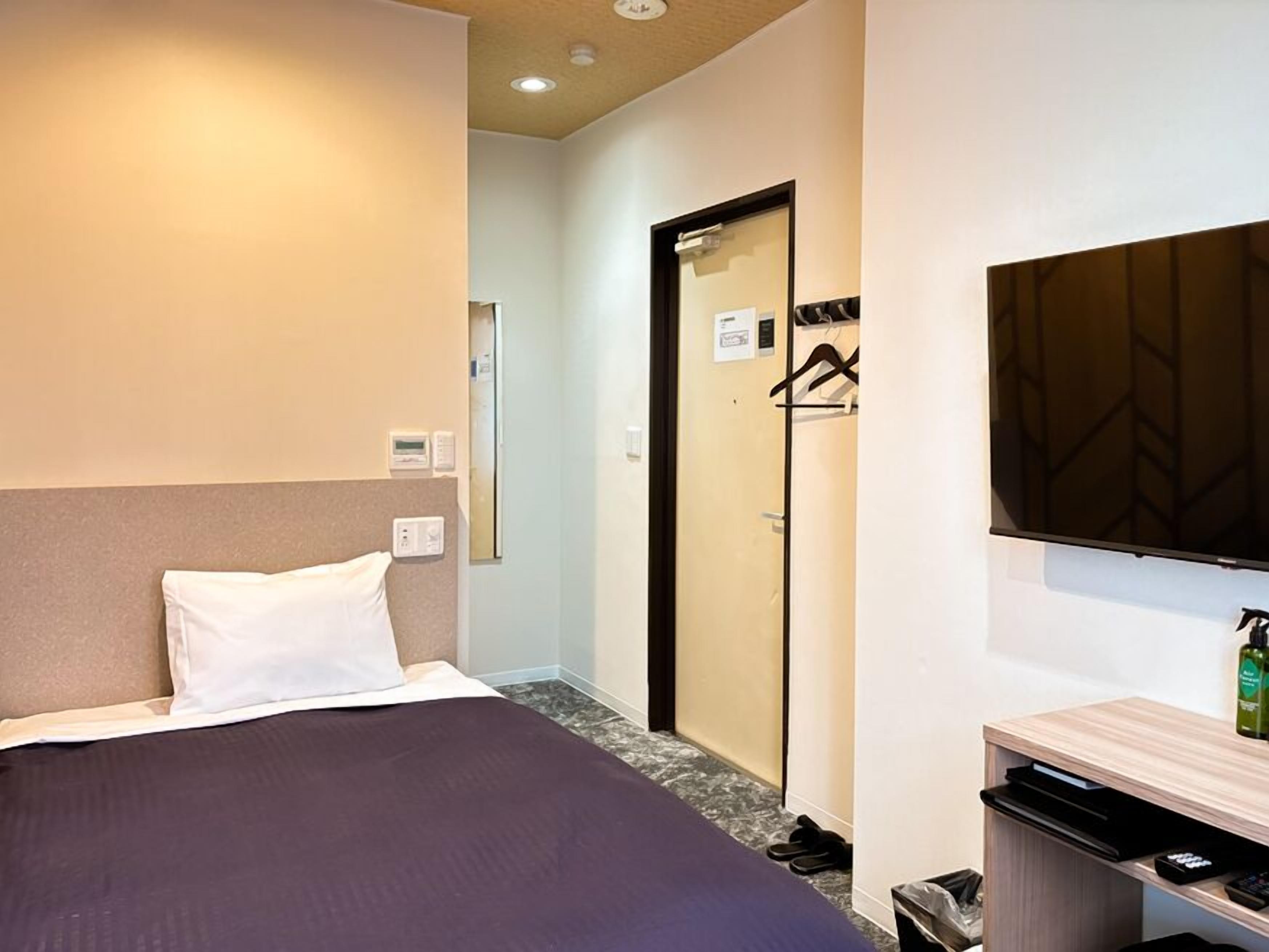 A hotel room with a bed, a TV, a mirror, a door, and a small desk with a bottle.