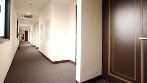 A hotel corridor with a carpeted floor, a wooden door with a gold frame, and a window on the left. - Hotel Livemax Kita Fuchu (Fuchu)
