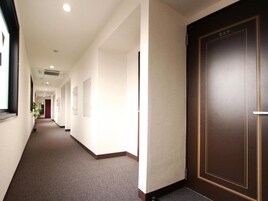 A hotel corridor with a carpeted floor, a wooden door with a gold frame, and a window on the left.