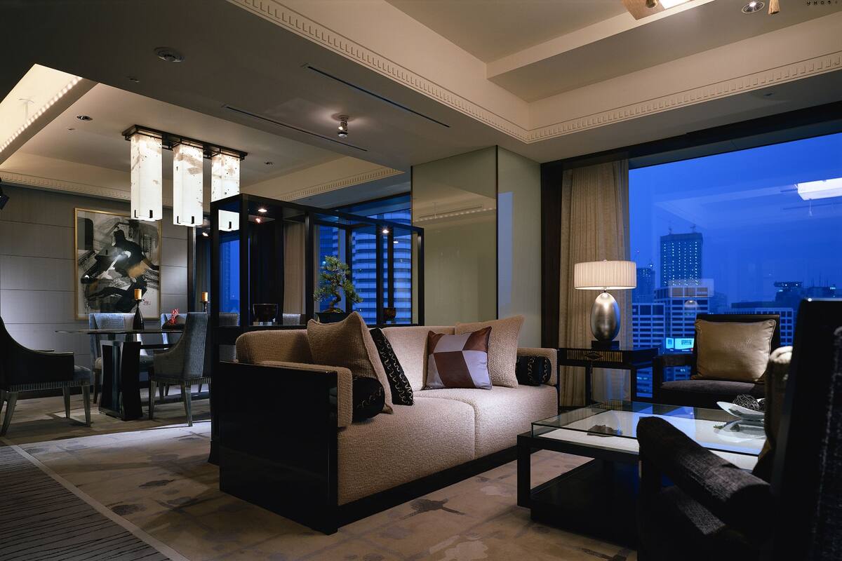 A modern hotel room with a large window offering a city view, a sofa, a dining table, and a separate seating area.