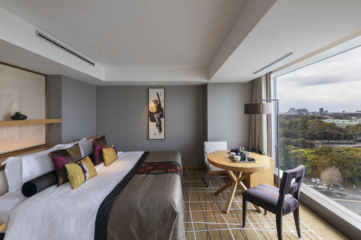 A modern hotel room with a large bed, a dining table, and a city view through the window.