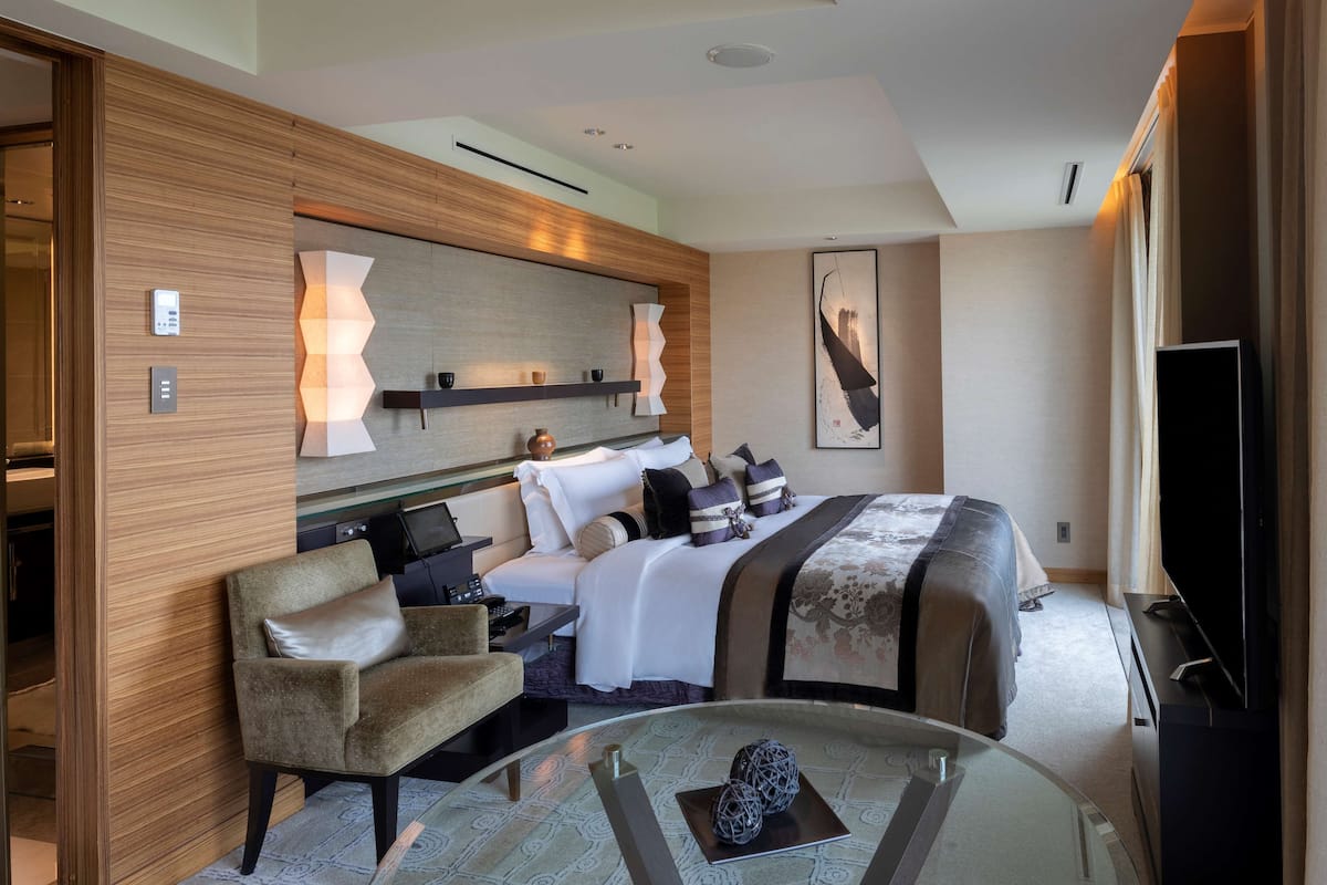 A modern hotel room with a bed, a seating area with a chair, a glass coffee table, and a flat-screen TV.