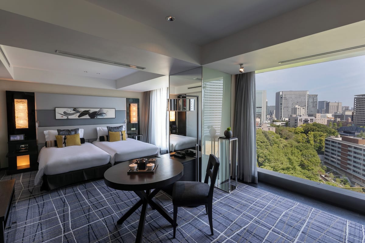 A modern hotel room with a city view, two beds, a dining table, and a large window.