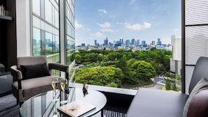 Habitación de hotel moderna con un amplio ventanal que ofrece vistas al horizonte de la ciudad, una zona de estar acogedora y una mesa de cristal con una bebida.