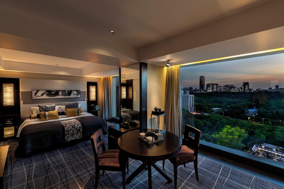 A modern hotel room with a large bed, a dining table, and a city view.