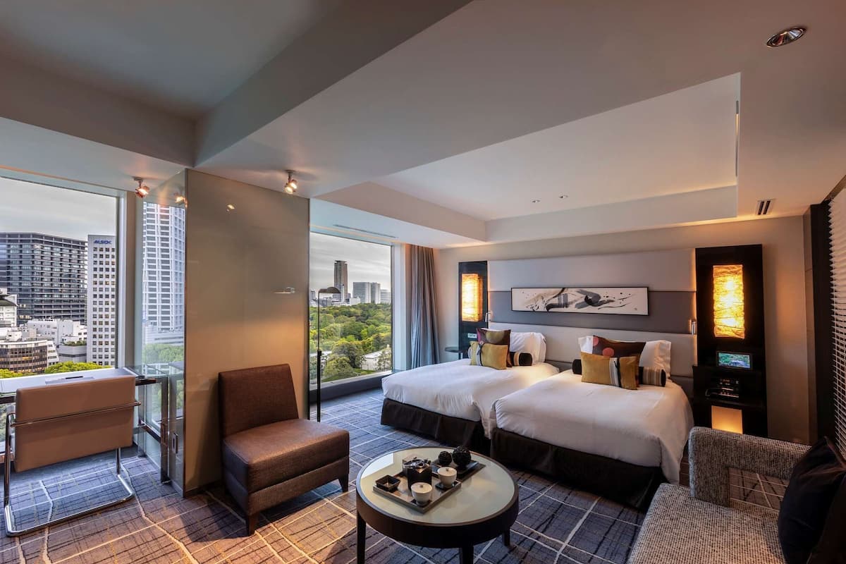 A modern hotel room with two beds, a desk, and a view of the city.
