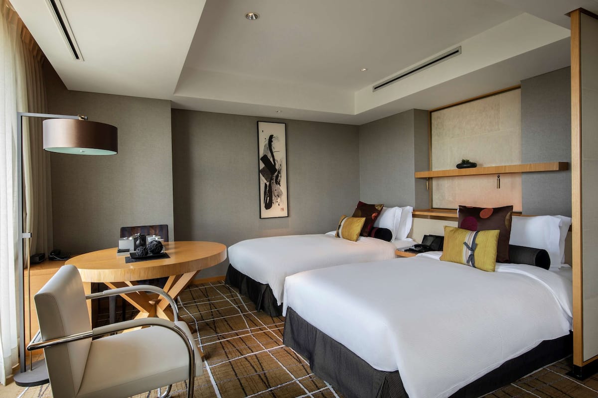 A hotel room with two beds, a round table, a chair, and a large window.