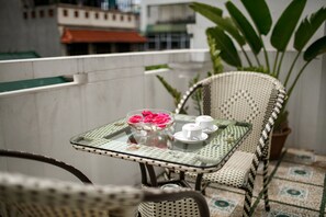 Una mesa con cubierta de vidrio, una silla trenzada en blanco y negro, un cuenco con flores rosadas y dos tazas blancas sobre un mantel estampado.