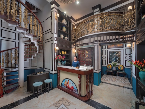 A reception area with a receptionist, a travel desk, and a reception counter. There is a staircase with ornate railings and a chandelier. - Hanoi Chic Boutique Hotel (Hanoi)