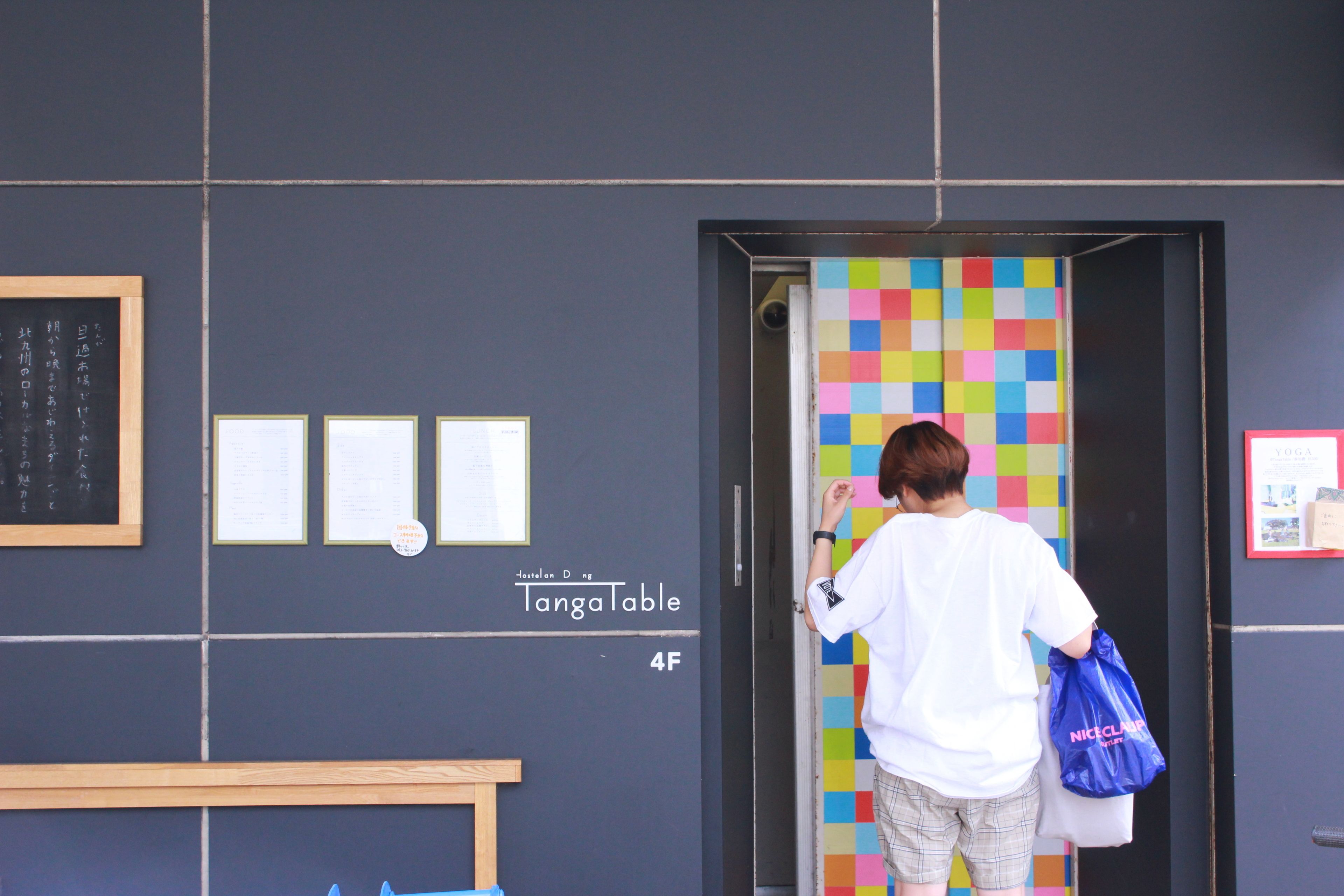 A person entering a room with a colorful wall, a wooden bench, and a sign for 'TangaTable' on the wall.