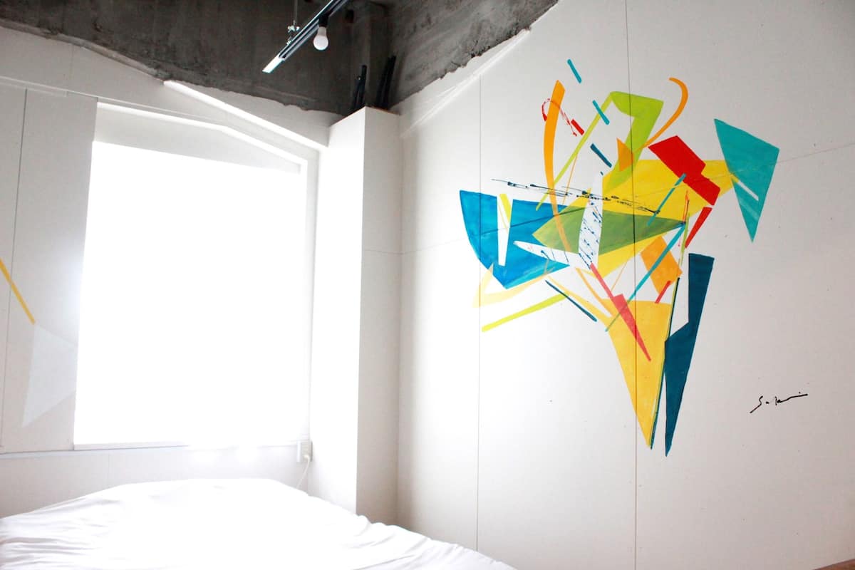 Ein Zimmer mit einem Bett, einem großen Fenster und einem farbenfrohen, abstrakten Wandbild.