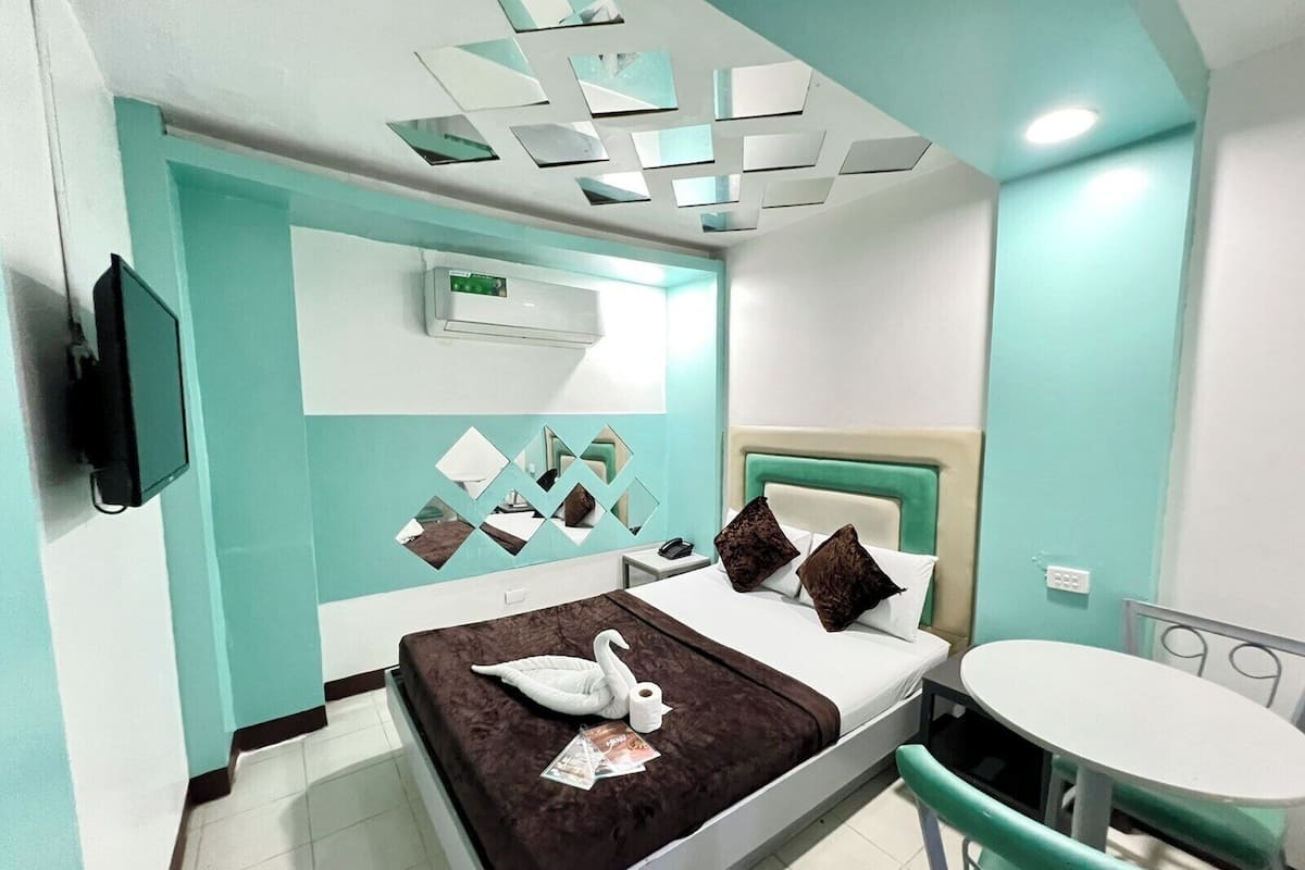 Habitación de hotel con cama, una mesita, una silla, televisor y aire acondicionado instalado en la pared.