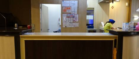 A hotel reception area with a wooden reception desk, a bulletin board with various notices, and a staff member in uniform.
