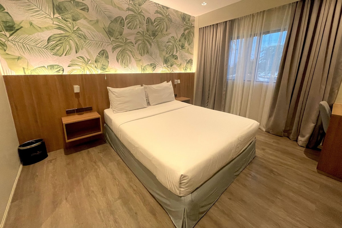 Habitación de hotel con una cama grande, cabecera de madera, una mesita de noche, una pequeña mesa y una ventana con cortinas.