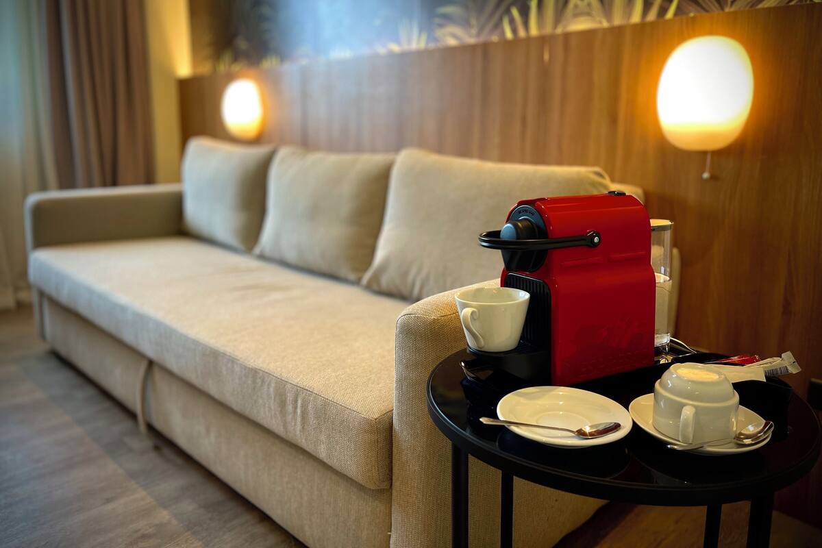 Habitación de hotel con sofá, cafetera, una taza, un plato y una cuchara sobre una mesita.