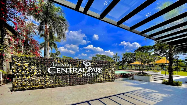 La entrada del Hotel Luisita Central Park, con un cartel grande, un camino empedrado y follaje circundante.