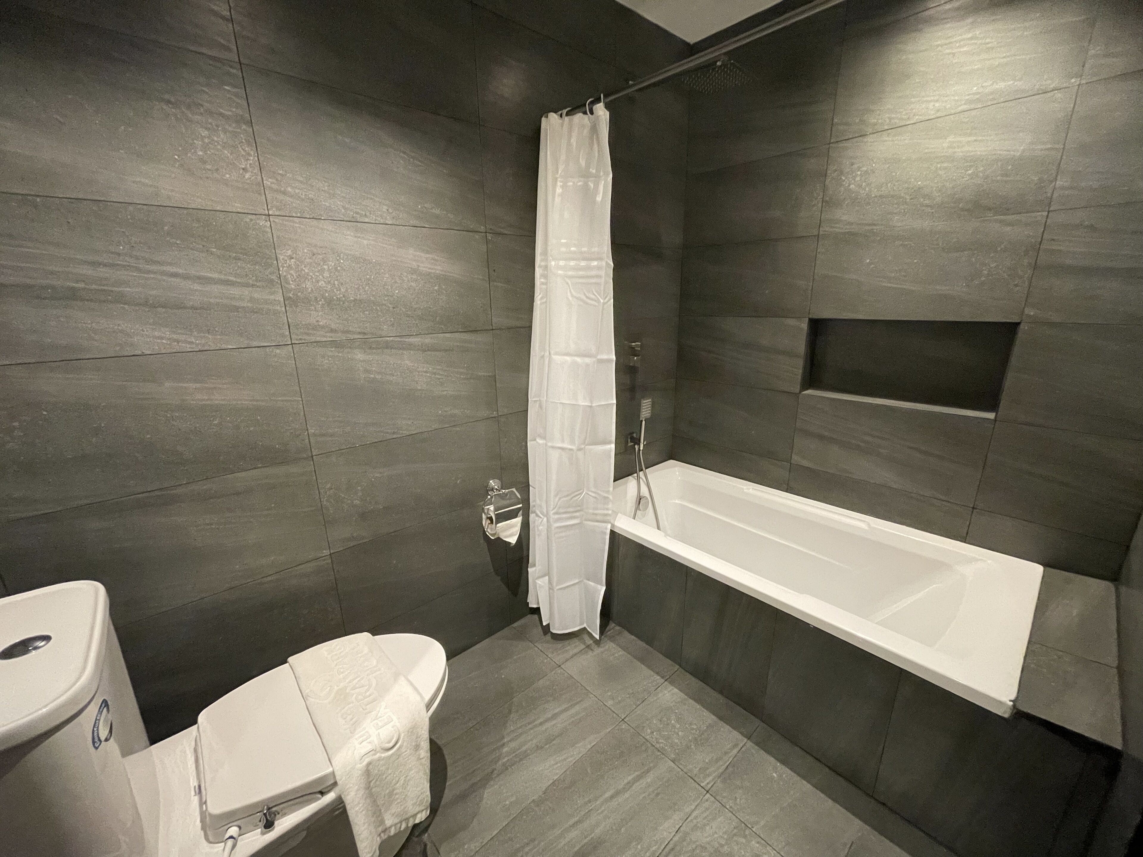 A modern bathroom with dark tiled walls, a white bathtub, and a toilet with a towel.